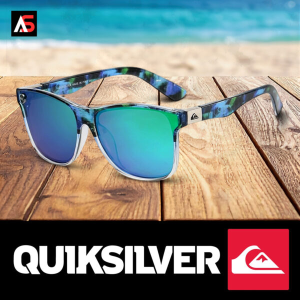 Angled lifestyle view of the Quiksilver 'Hydro-Camo' Square sunglasses with a clear frame and ombre blue mirror lens, set on a beach with the ocean waves in the background.